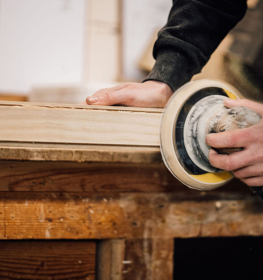 A close-up of a yellow hand sander, finishing the edges of a large timber frame on a workbench. Tucker Joinery, Andover.