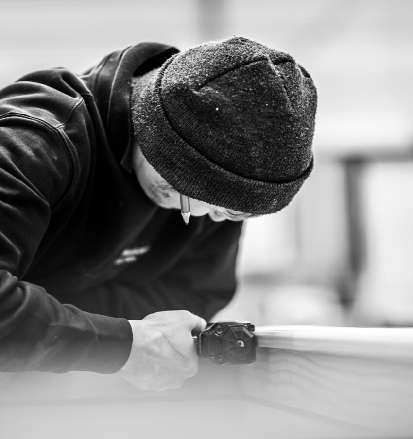 A member of the Tucker Joinery team works on the trim of a large timber frame with a hand tool. The man leans in closely to his work at the wood workshop based in Andover.