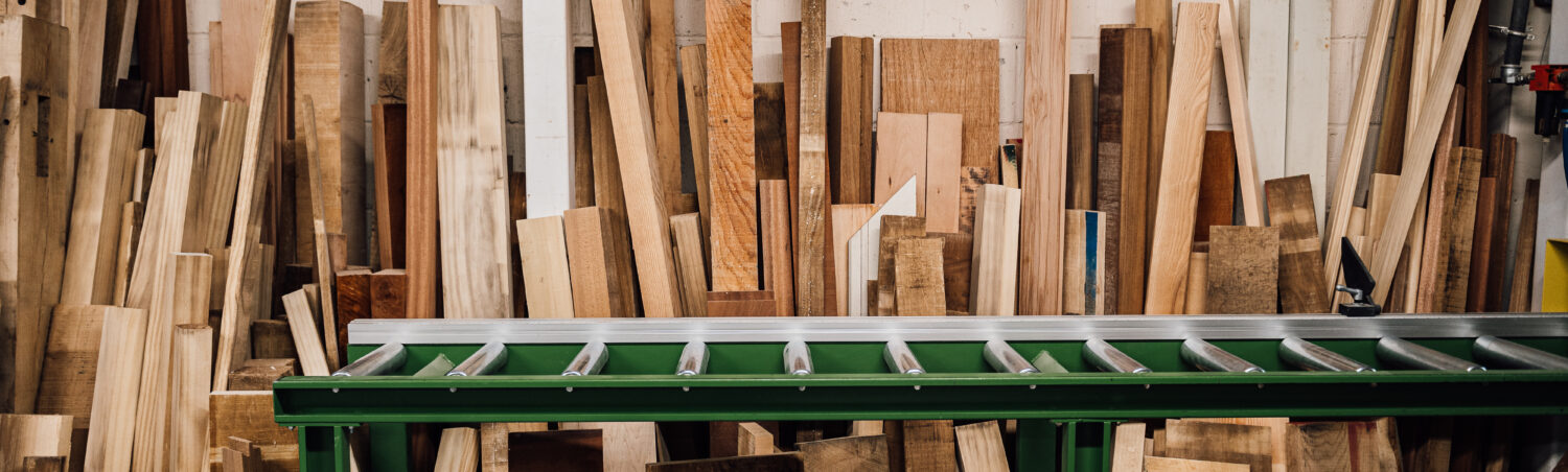 A range of cut timber is stood against the back wall of the Tucker Joinery workshop. A green ladder sits atop a workbench in the foreground.