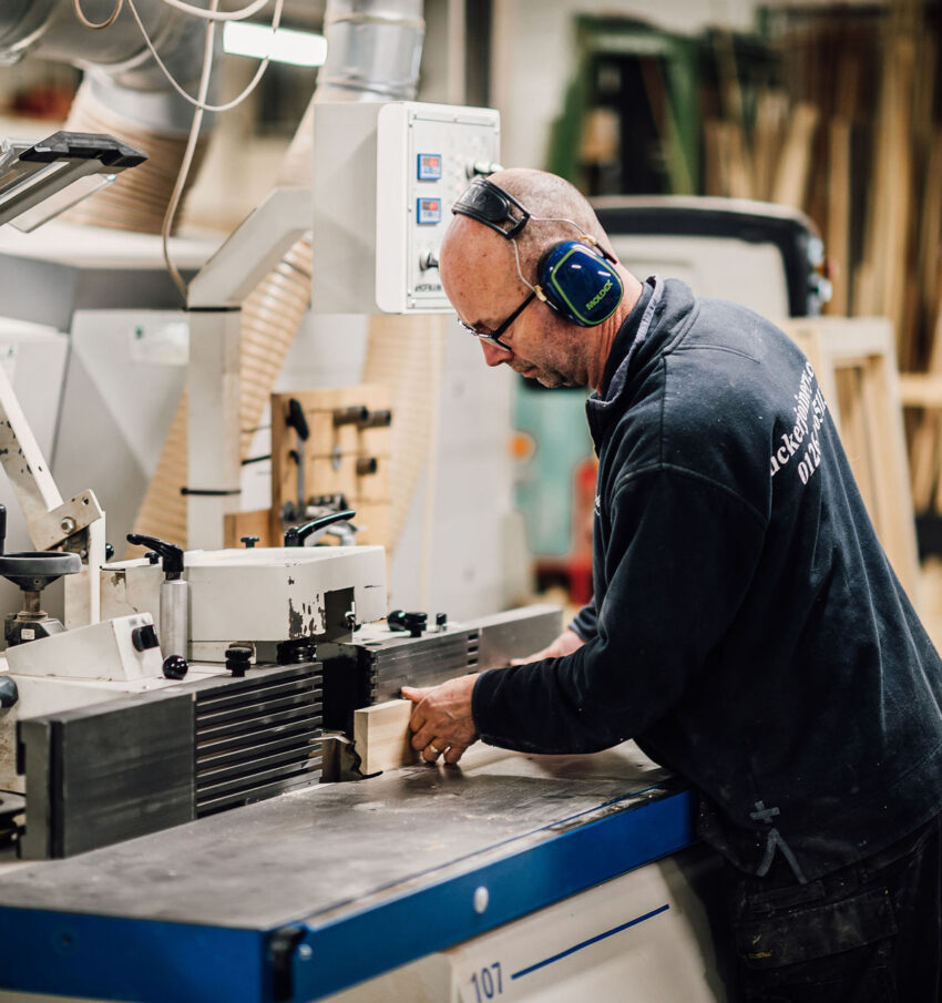 A member of the Tucker Joinery team in safety gear and a branded sweatshirt, shapes timber mouldings in a machine at the workshop. Andover.